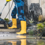 Men Wearing Yellow Rubber Boots Pressure Washing Architectural Concrete Elements in His Garden. Lower Body Close Up. Modern Concrete Backyard Stairs Cleaning.